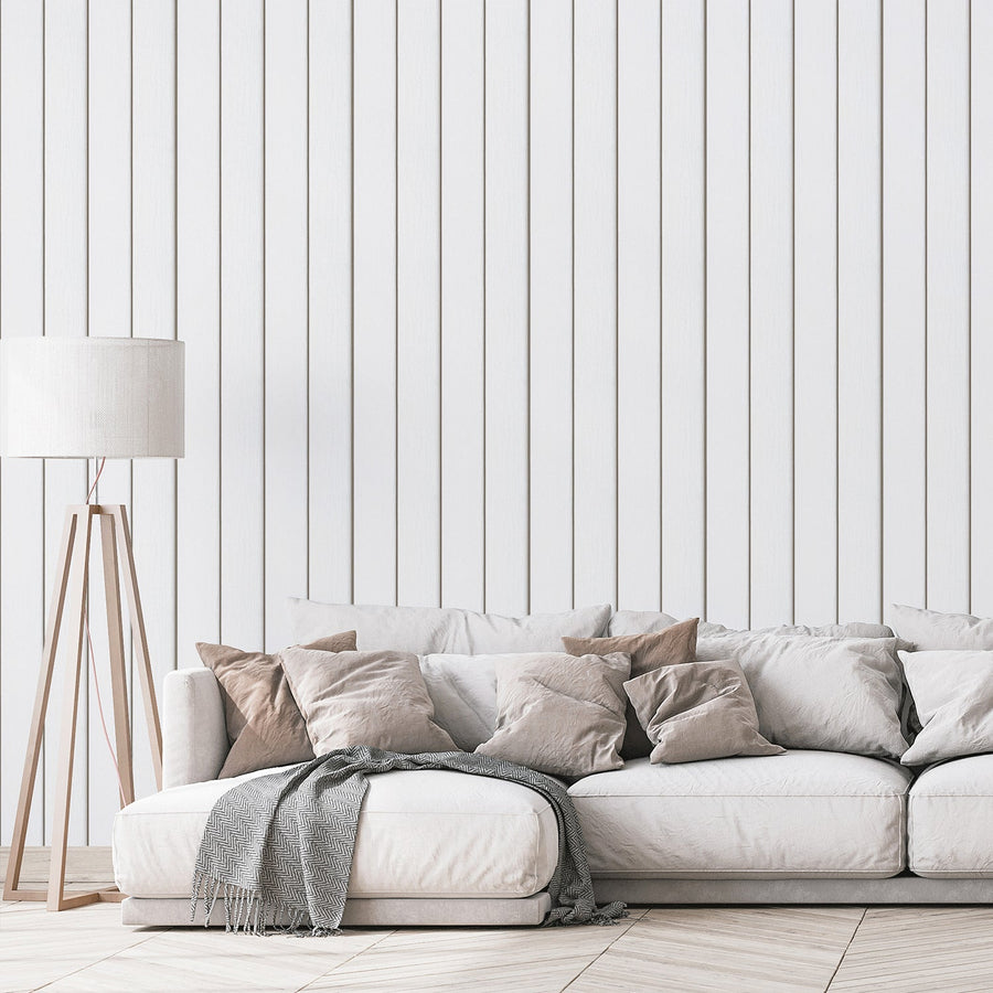 Ett vardagsrum med en golvlampa och en soffa i neutral färg, med tapeten 'Airin', en vit tapet med vertikala träplankor med realistisk ådring och subtil väderbiten struktur i en rustik paneldesign.