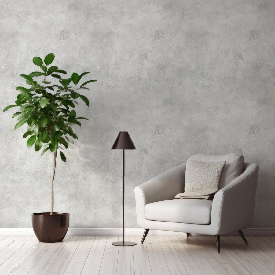 Ett vardagsrum med en grå fåtölj bredvid en golvlampa och en bladväxt samt tapeten Ash, en grå tapet med betongeffekt, detaljerad stenliknande struktur och mjuka tonvariationer för en industriell look.