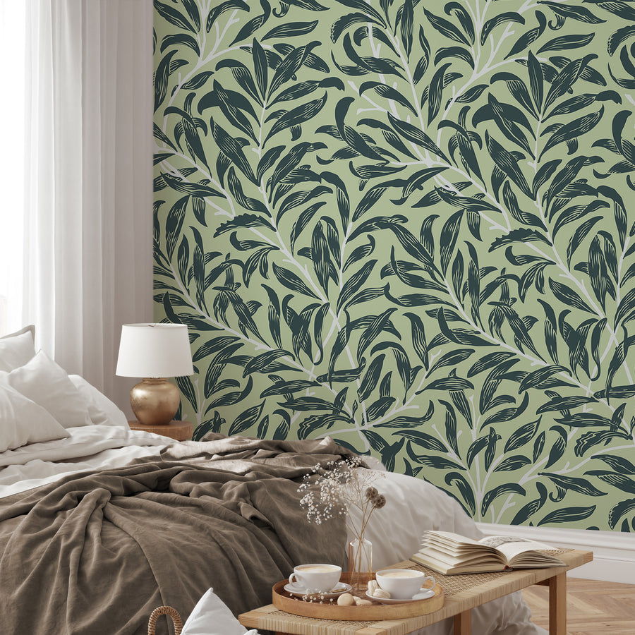 Ett sovrum i neutral stil med sängkläder, lampa och en bänk vid fotänden och Lyra, en botanisk tapet med skiktade gröna tropiska blad och sammanflätade lummiga rankor på mjuk neutral bakgrund i ett frodigt upprepat bladmönster.