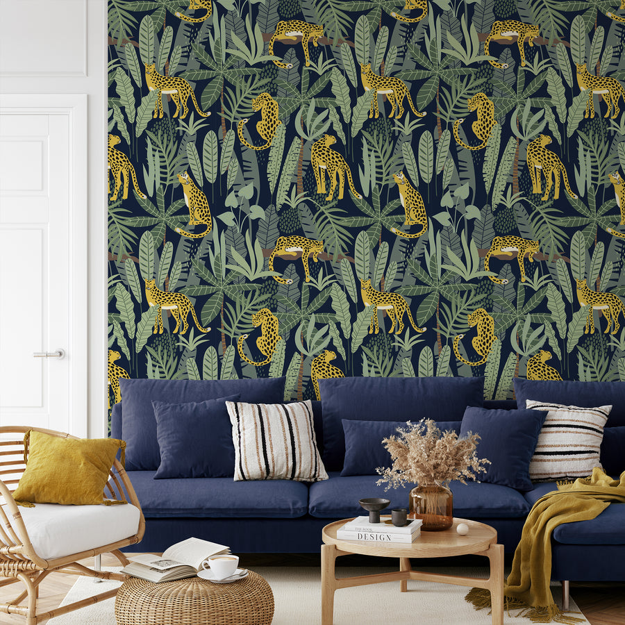 Ett vardagsrum med en marinblå soffa full av kuddar och ett lågt soffbord, med tapeten 'Clymene', en safaritapet med illustrerade geparder bland frodiga tropiska palmblad och exotiskt grönt bladverk mot en mjuk neutral bakgrund i ett upprepande djungelmönster.