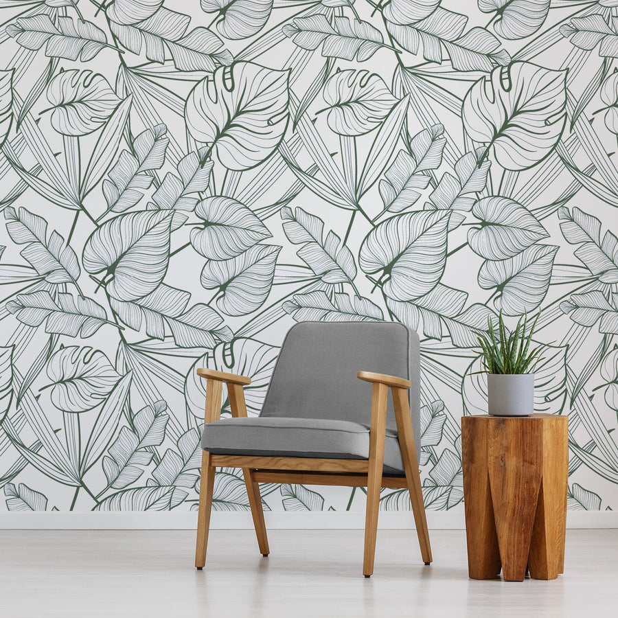 Ett vardagsrum med en grå fåtölj och ett litet sidobord med en växt på, med tapeten 'Juniper Dark Green', en tropisk tapet med mörkgröna konturer av palmblad och frodigt botaniskt bladverk mot en krispigt vit bakgrund i ett djärvt upprepat mönster.