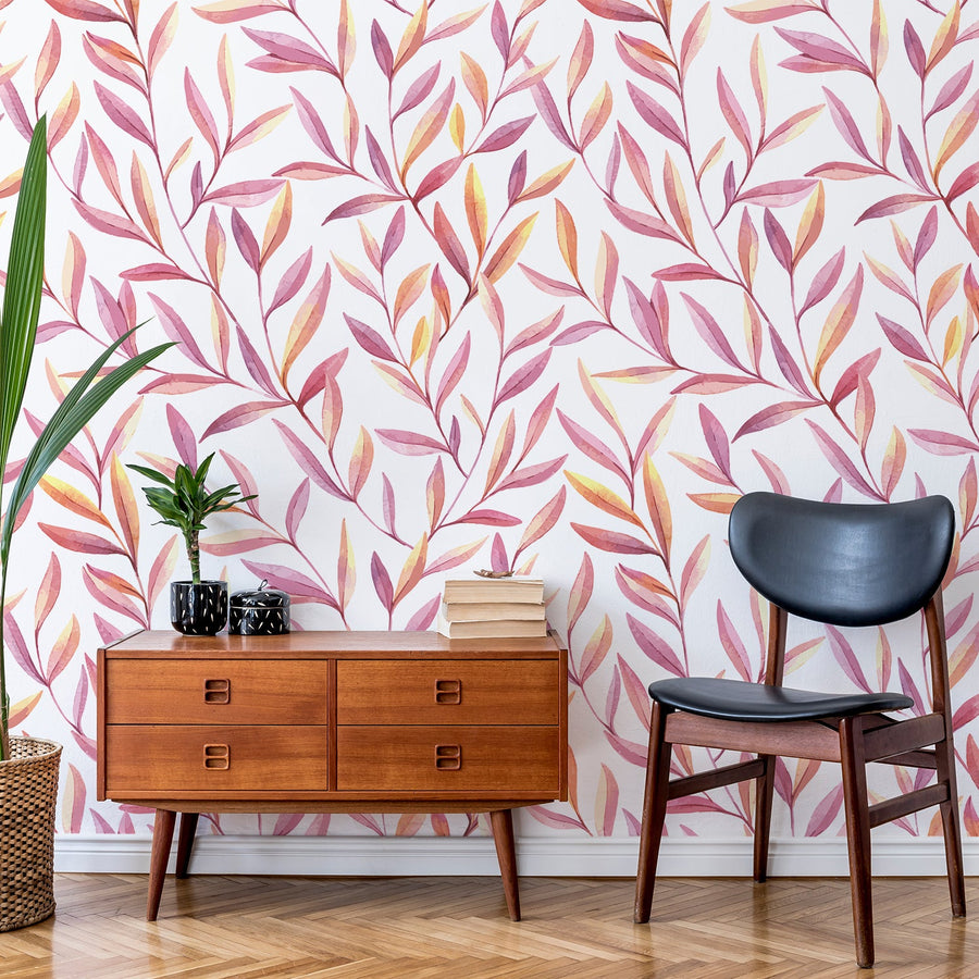 Ett vardagsrum med en svart läderfåtölj och en byrå i bärnstensfärgat trä med lummiga gröna växter, med tapeten 'Perry', en botanisk tapet med rosa och orange akvarellblad mot en krispigt vit bakgrund i ett mjukt, modernt upprepat bladmönster.