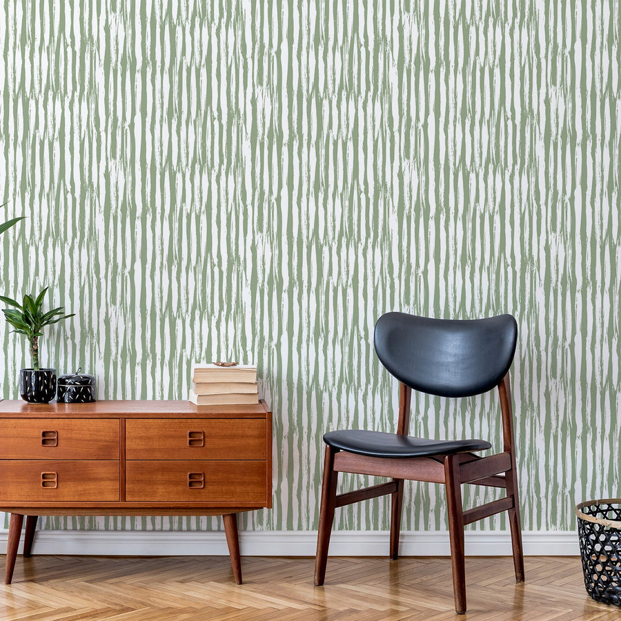 Ett vardagsrum med en svart läderfåtölj bredvid ett bärnstensfärgat sideboardskåp, med tapeten 'Piper', en grön och vit tapet med tunna vertikala linjer i ett enkelt randigt mönster med en ren, modern geometrisk design.