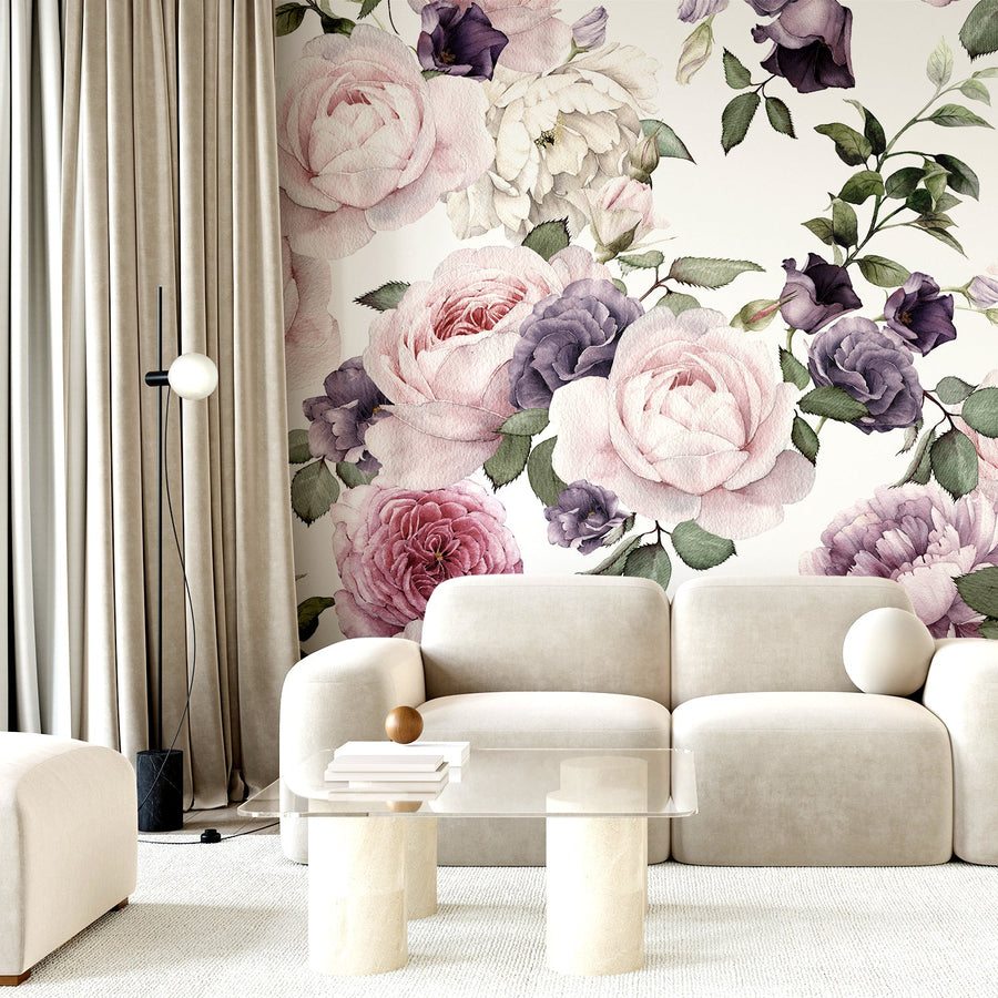Ett vardagsrum med en krämvit soffa, ett soffbord i glas och mjuka gardiner, med tapeten 'Rosalie', en rosa och lila tapet med mjuka akvarellrosor och fina blomdetaljer i ett romantiskt upprepat botaniskt mönster.